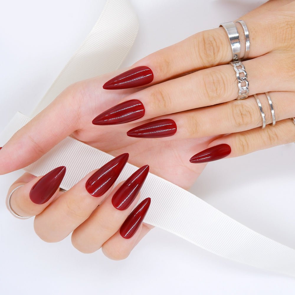 Hands showing fingers adorned in long stilletto shaped maroon color press-on nails,holding a white ribbon against a white background.