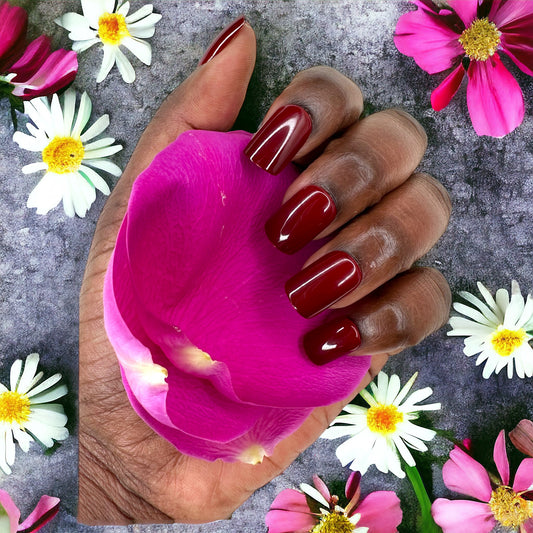 Hand holding pink flower petals with maroon short square press-on nails against a pink and white flowered background