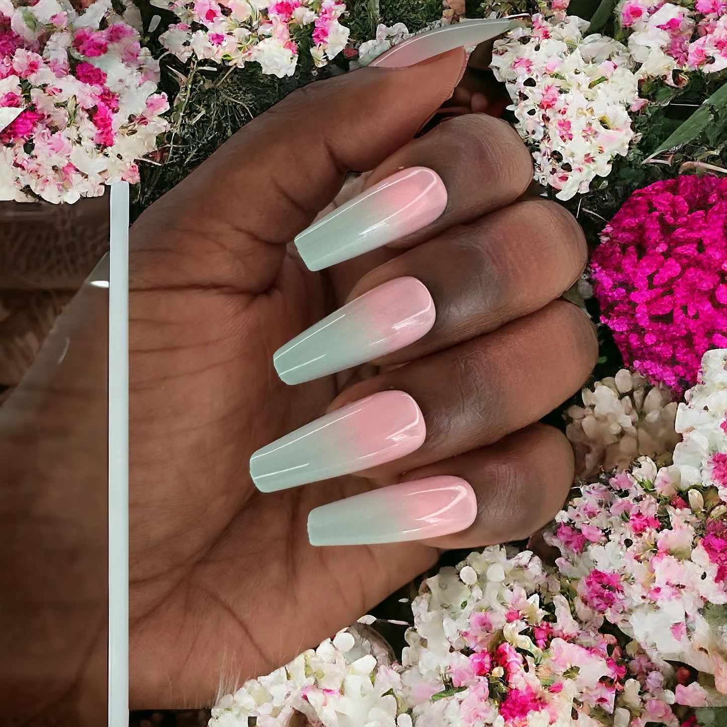 Fingers with pink and blue ombre color long coffin shape press-on nails against a backdrop of pink and white flowers