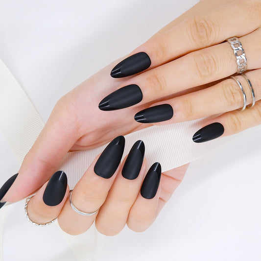 Two hands with medium almond-shaped press-on nails in black matte and glossy tips, one hand holding a white ribbon against a white background