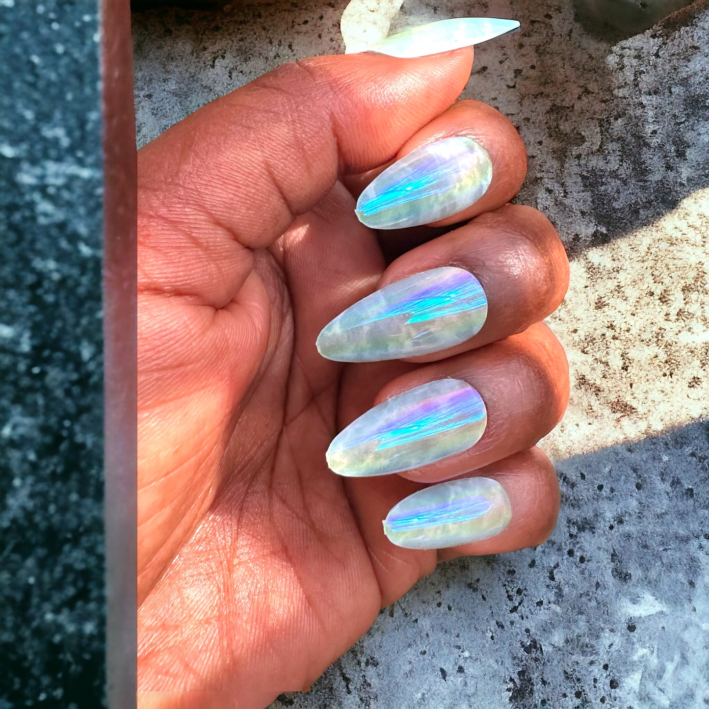 A hand with almond-shaped press-on nails featuring a sky blue marble design, set against a grey concrete background