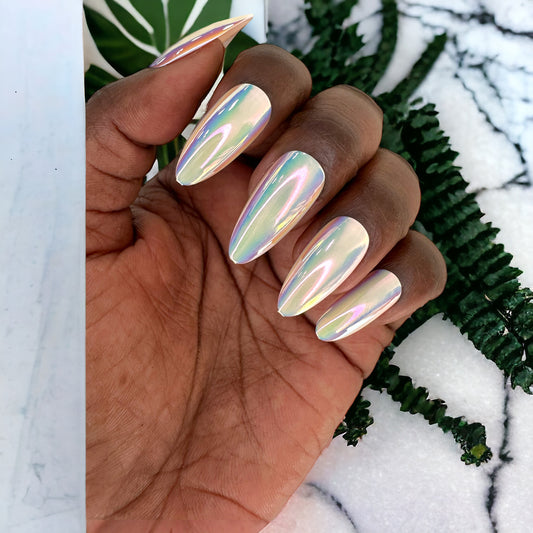 A hand displaying almond-shaped press-on nails in white chrome, set against a background of lush green plants