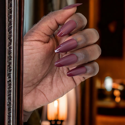 A hand featuring almond-shaped press-on nails in matte maroon with glossy tips, set against a warm, softly lit background