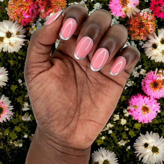 A hand with short round press-on nails featuring pink hues and silver tips, set against a colorful background of flowers